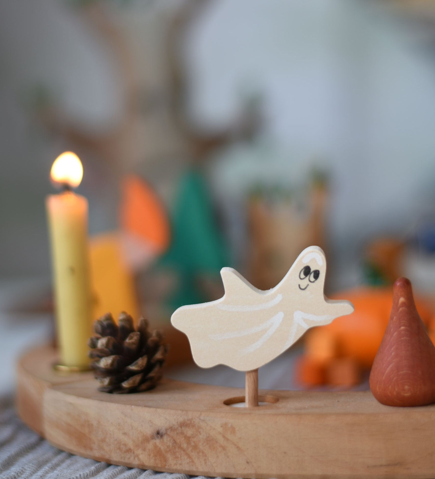 A Grimm's spooky ghost decorative figure placed on a natural celebration ring. A lit beeswax candle and other decorative figures can be seen in the background. The decorative figures are part of a wide range of Waldorf inspired figures and celebration rings available here at Babipur.