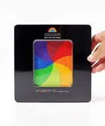 An adult hand holding a closed Grimms tin with a clear window in the front showing the Goethe magnetic puzzle set