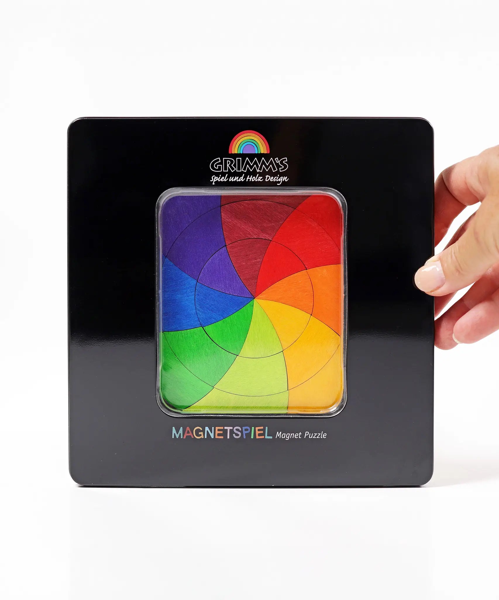 An adult hand holding a closed Grimms tin with a clear window in the front showing the Goethe magnetic puzzle set