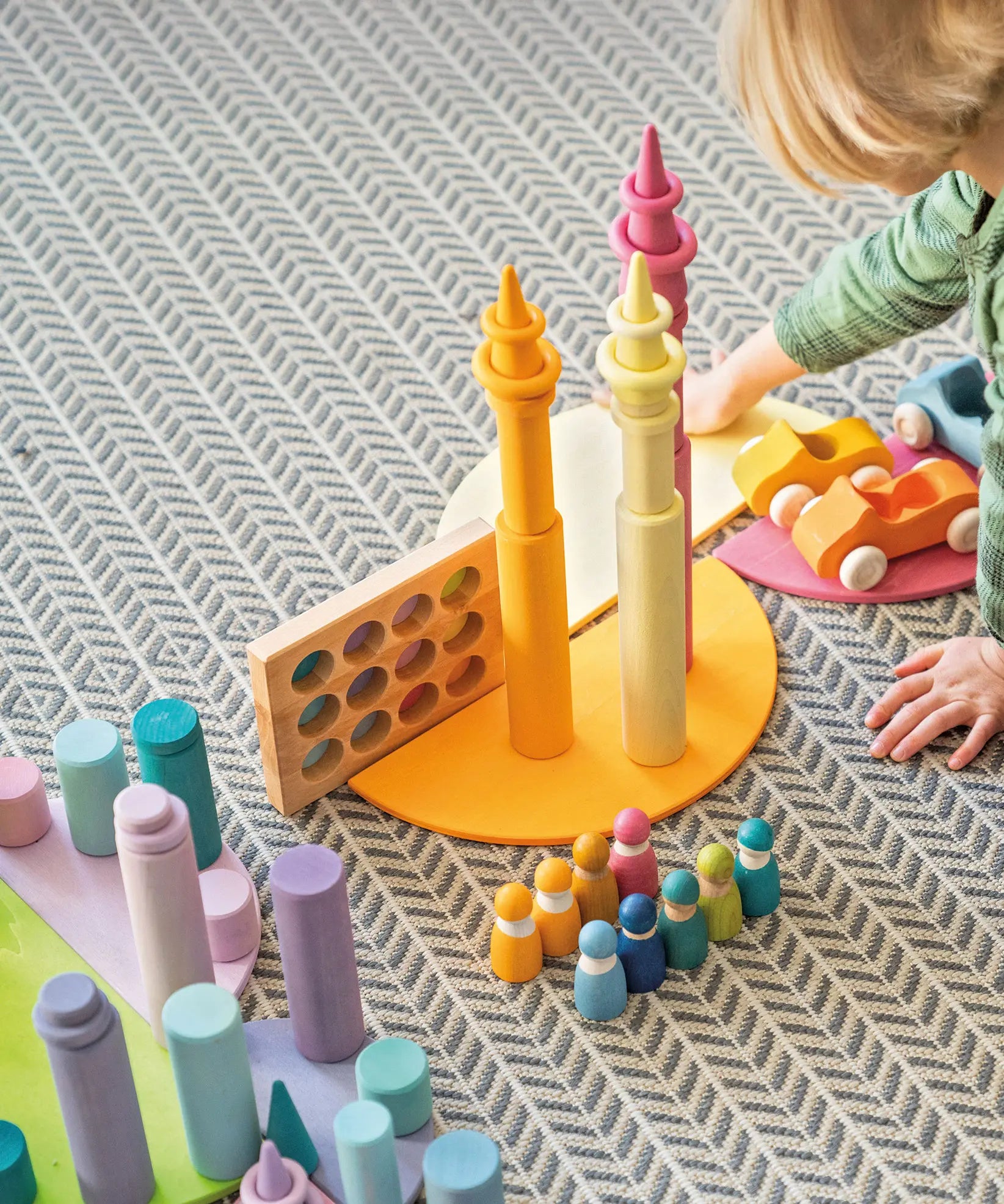 Child playing with Grimm's wooden open ended toys including large building rollers in pastel, friends peg dolls, forest pieces and cars.