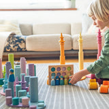 Child playing with Grimm's wooden open ended toys including large building rollers in pastel, friends peg dolls, forest pieces and cars.