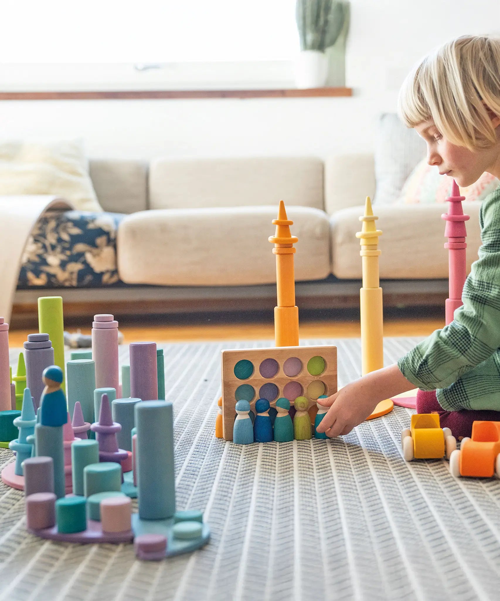 Child playing with Grimm's wooden open ended toys including large building rollers in pastel, friends peg dolls, forest pieces and cars.
