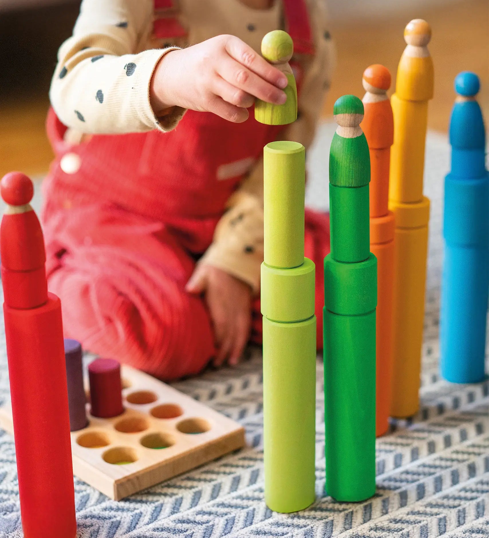 Child playing with the Grimm's large building rollers in bright rainbow colours stacking friends peg dolls on top. 