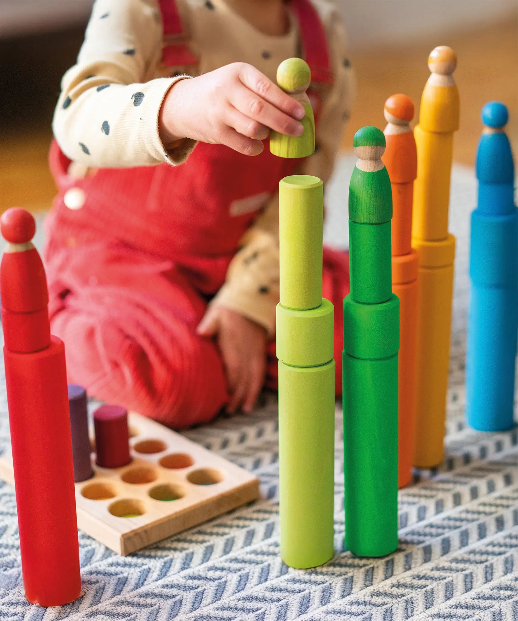 Child playing with the Grimm's large building rollers in bright rainbow colours stacking friends peg dolls on top. 