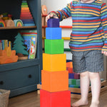 A child playing with the Grimm's Large Coloured Boxes Set. The boxes are all stacked up on top of each other, the child is placing the smallest block on the top