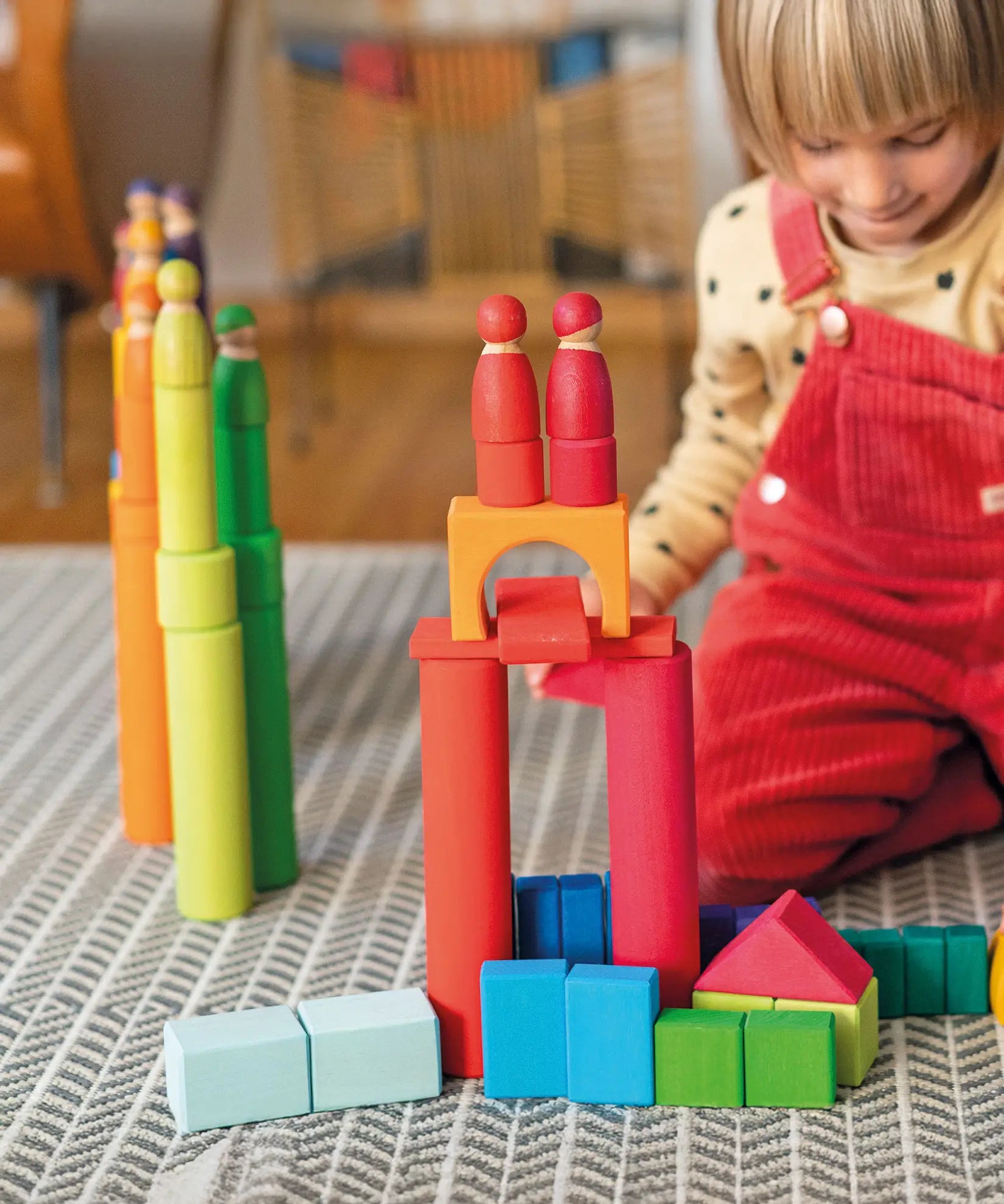 Child playing with the Grimm's large building rollers in bright rainbow colours with wooden blocks and friends peg dolls.