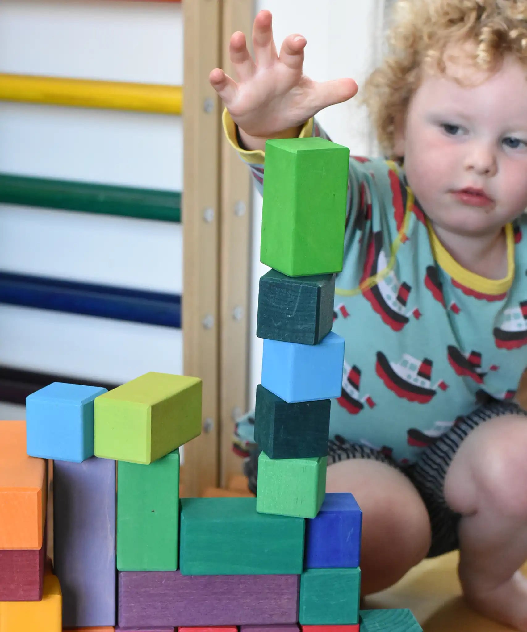 Grimm's large stepped pyramid coloured wooden block set played with in the Babipur playroom. Close up of child's hand reaching for a green block.