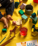 Loose hand carved tactile pieces from the Grimms movement building set on a yellow surface next to child's feet