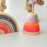 Child hand holding a small wooden Grimms conical piece from the neon pink tower next to matching arched pieces showing scale