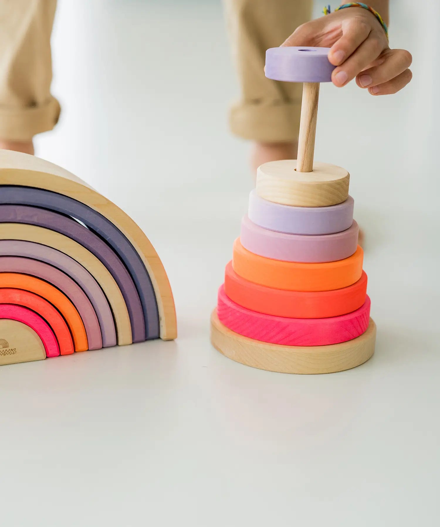 Child hand holding a small wooden Grimms conical piece from the neon pink tower next to matching arched pieces showing scale