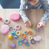 Top view of a child holding a pile of Grimms wooden pastel building rings on a table next to wooden pink pebble pieces