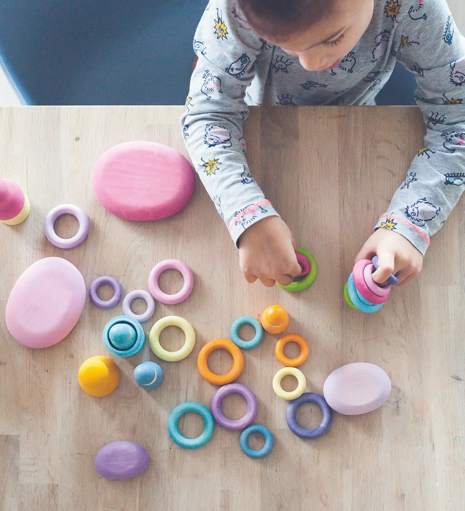 Top view of a child holding a pile of Grimms wooden pastel building rings on a table next to wooden pink pebble pieces