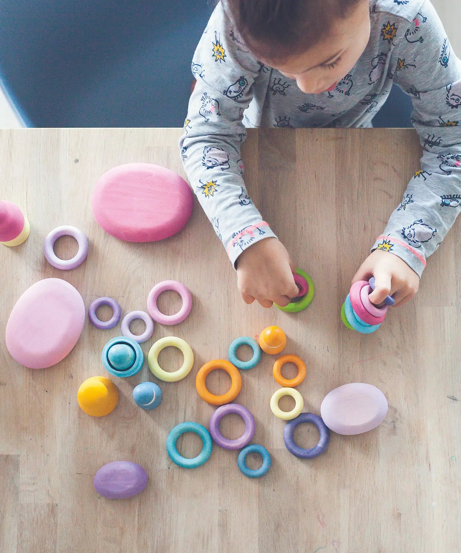 Top view of a child holding a pile of Grimms wooden pastel building rings on a table next to wooden pink pebble pieces
