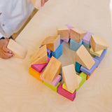 Child holding a triangular natural wooden block next to a structure made from the Grimms pastel duo wooden block set