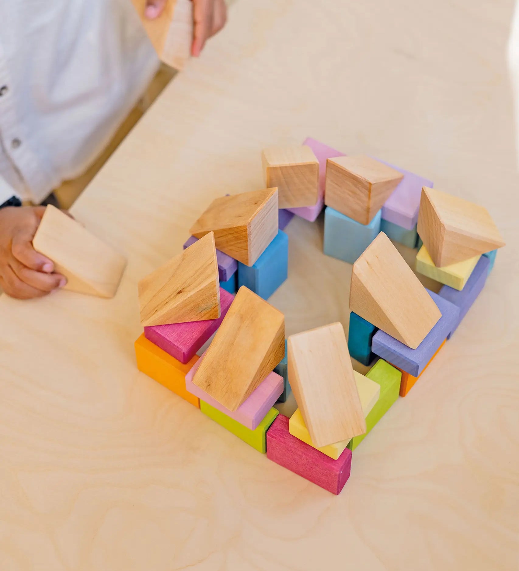 Child holding a triangular natural wooden block next to a structure made from the Grimms pastel duo wooden block set