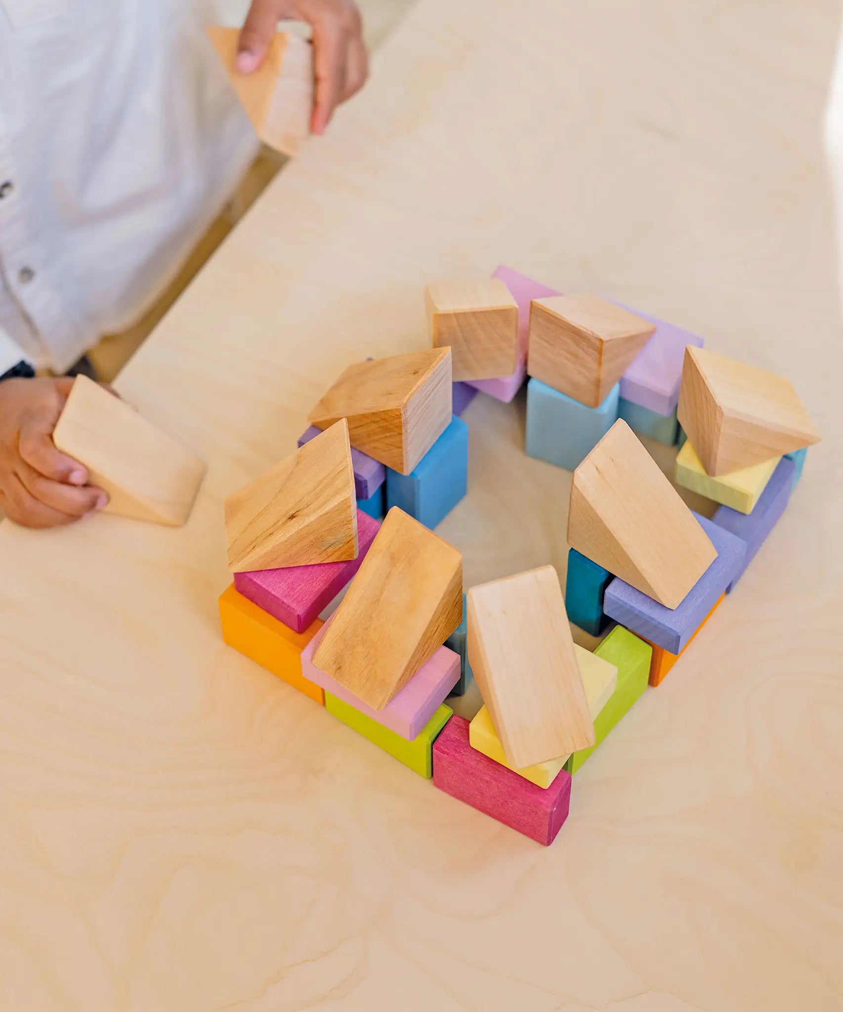 Child holding a triangular natural wooden block next to a structure made from the Grimms pastel duo wooden block set