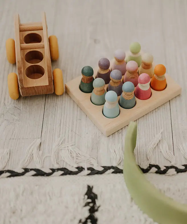 Grimms handcrafted sorting board with pastel coloured peg dolls sitting inside the round slots next to a toy bus 