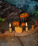A Grimm's pumpkin decorative figure placed on a natural Grimm's celebration ring in between two lit candles. Bumbu Halloween pumpkins can be seen in the background. 