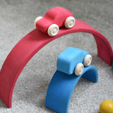 The red and blue cars from the Grimm's 6 Coloured Wooden Cars set placed on matching coloured pieces from the large Grimm's rainbow tunnel. These wooden toy cars are part of a wide range of Grimm's wooden toys available here at Babipur.
