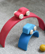 The red and blue cars from the Grimm's 6 Coloured Wooden Cars set placed on matching coloured pieces from the large Grimm's rainbow tunnel. These wooden toy cars are part of a wide range of Grimm's wooden toys available here at Babipur.
