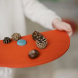 Child holing a Grimms wooden red semi circle piece with acorns and pine cones on top