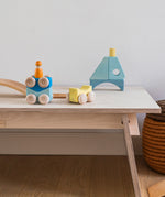 Grimm's wooden stacking car toys placed on a wooden table with a Grimm;s friend peg doll on top of them. These Grimm's toys are part of a wide range of wooden toys available here at Babipur. 