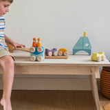 A child playing with the Grimm's wooden stacking car toys placed on a wooden table with a Grimm's friend peg doll on top of them. These Grimm's toys are part of a wide range of wooden toys available here at Babipur. 
