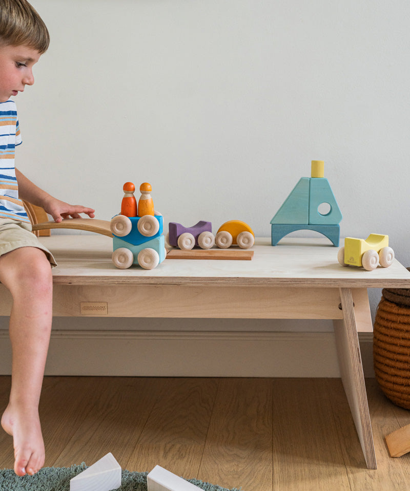 A child playing with the Grimm's wooden stacking car toys placed on a wooden table with a Grimm's friend peg doll on top of them. These Grimm's toys are part of a wide range of wooden toys available here at Babipur. 