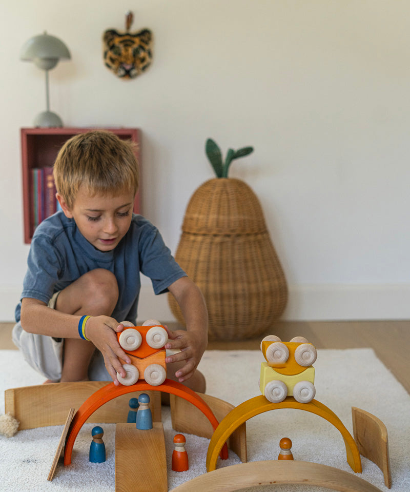 A child playing with the orange Grimm's wooden stacking car toys placing them on a Grimm's bridge piece. These Grimm's toys are part of a wide range of wooden toys available here at Babipur. 