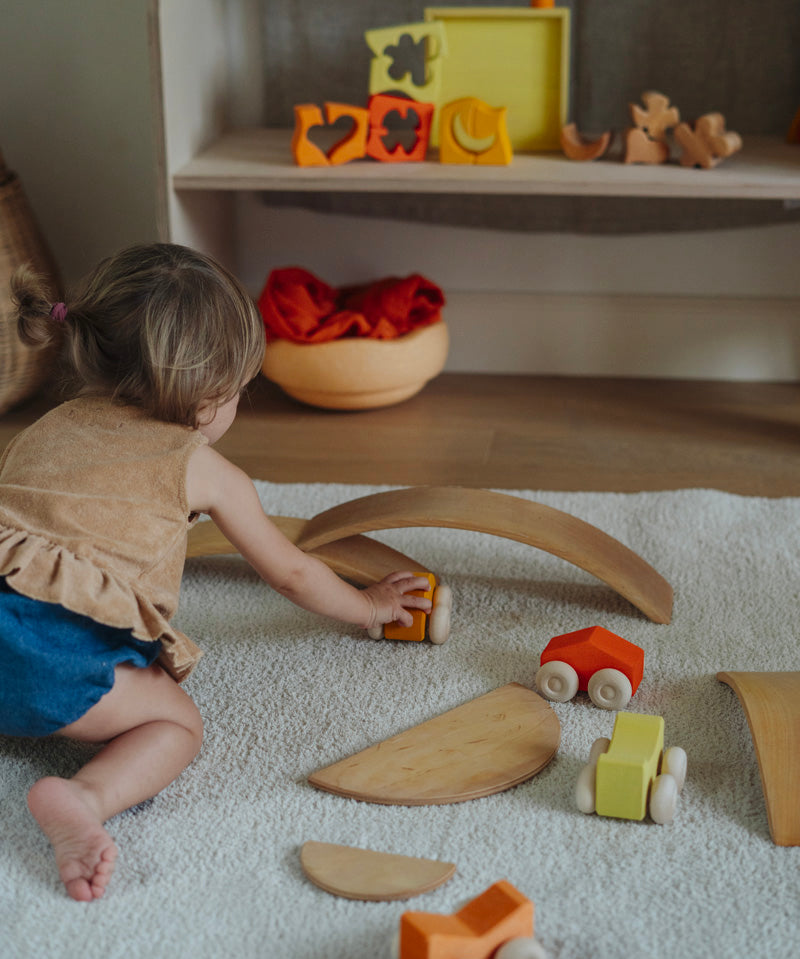 A child playing with the orange Grimm's wooden stacking car toys driving them under a Grimm's bridge piece. These Grimm's toys are part of a wide range of wooden toys available here at Babipur. 
