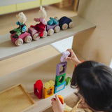 A line of purple and pink Grimm's wooden stacking car toys on a wooden shelf. A child is standing below playing with the Grimm's window set. These Grimm's toys are part of a wide range of wooden toys available here at Babipur. 