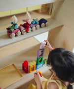A line of purple and pink Grimm's wooden stacking car toys on a wooden shelf. A child is standing below playing with the Grimm's window set. These Grimm's toys are part of a wide range of wooden toys available here at Babipur. 