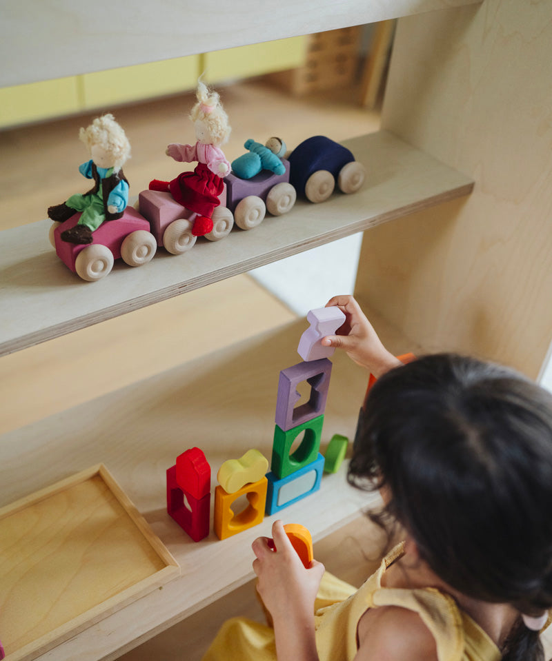 A line of purple and pink Grimm's wooden stacking car toys on a wooden shelf. A child is standing below playing with the Grimm's window set. These Grimm's toys are part of a wide range of wooden toys available here at Babipur. 