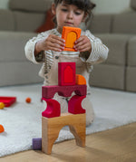 A child playing with the Grimm's red car stacking puzzle building block set stacking pieces from the Grimm's window set on top of a stack of the blocks. These Grimm's toys are part of a wide range of wooden toys available here at Babipur. 