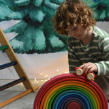 A child pushing a red coloured Grimm's wooden toy car over the large rainbow. A Ticlimb climbing frame can be seen in the background along with a Wonderie Christmas tree design cloth hung up on the wall behind. These Grimm's toys are part of a wide range of wooden toys available here at Babipur. 