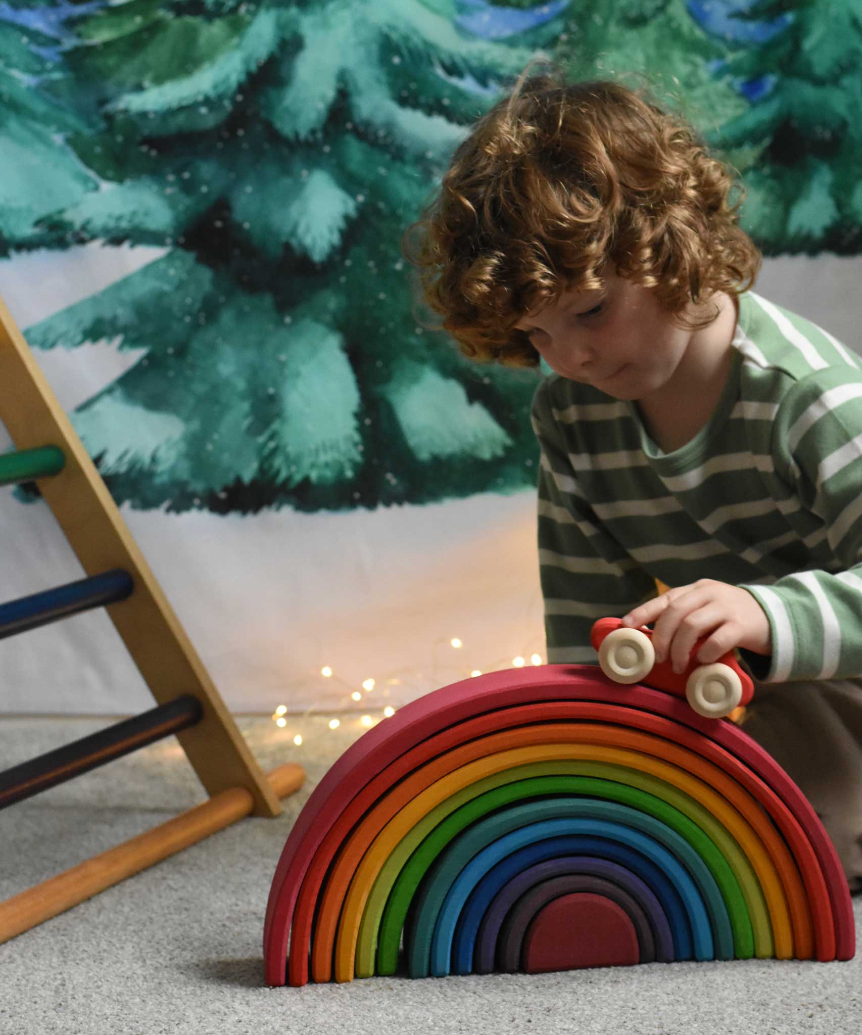 A child pushing a red coloured Grimm's wooden toy car over the large rainbow. A Ticlimb climbing frame can be seen in the background along with a Wonderie Christmas tree design cloth hung up on the wall behind. These Grimm's toys are part of a wide range of wooden toys available here at Babipur. 