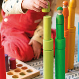 Child playing with the Grimm's large and small building rollers in a rainbow palette stacking friends peg dolls on top. 