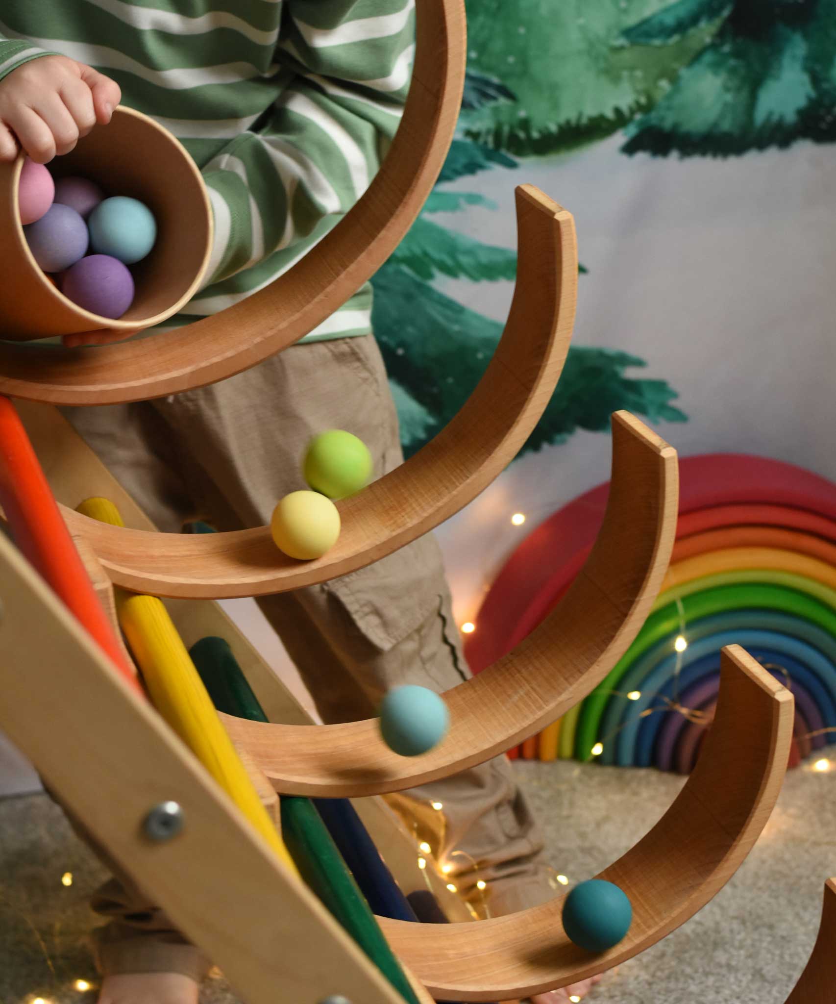 A child tipping out a tub of Grimm's small pastel balls onto a ball run created using the Grimm's natural rainbow and a Triclmb climbing frame.
These Grimm's toys are part of a wide range of wooden toys available here at Babipur.  