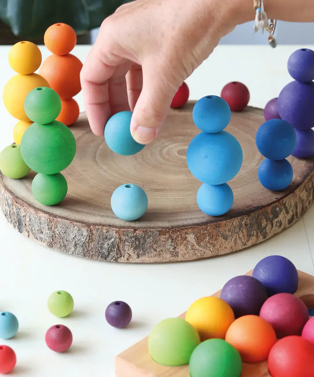 Adult hand holding a light blue Grimms wooden small ball next to other stacked balls on a wooden slab