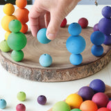 Adult hand holding a light blue Grimms wooden small ball next to other stacked balls on a wooden slab