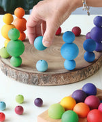 Adult hand holding a light blue Grimms wooden small ball next to other stacked balls on a wooden slab