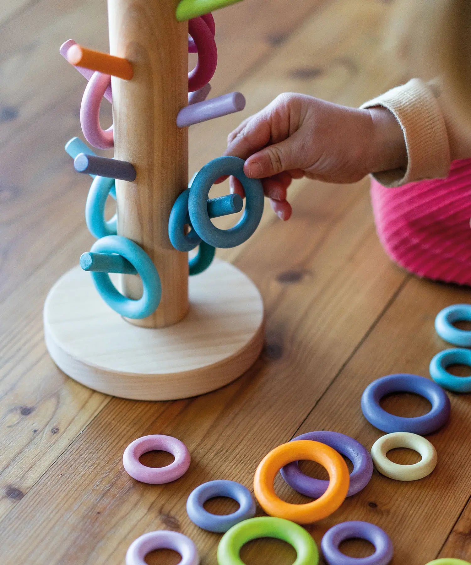 Grimms wooden sorting helper in pastel standing next to matching loose ring pieces and a child holding a blue ring