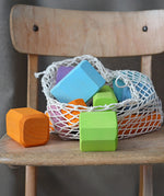 Grimm's tree slices wooden coloured building blocks shown in their netting storage bag placed on a wooden chair. 