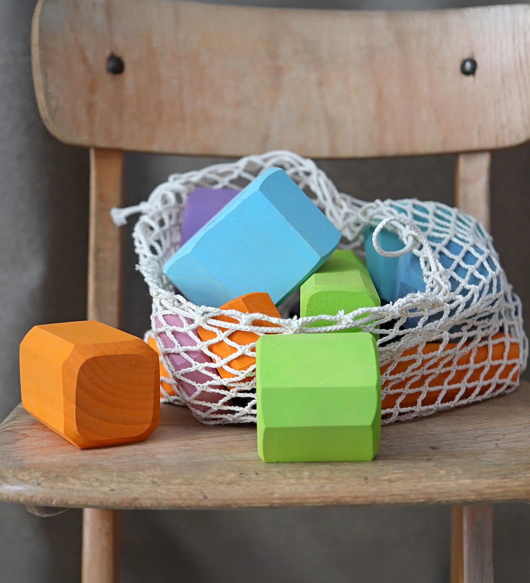 Grimm's tree slices wooden coloured building blocks shown in their netting storage bag placed on a wooden chair. 