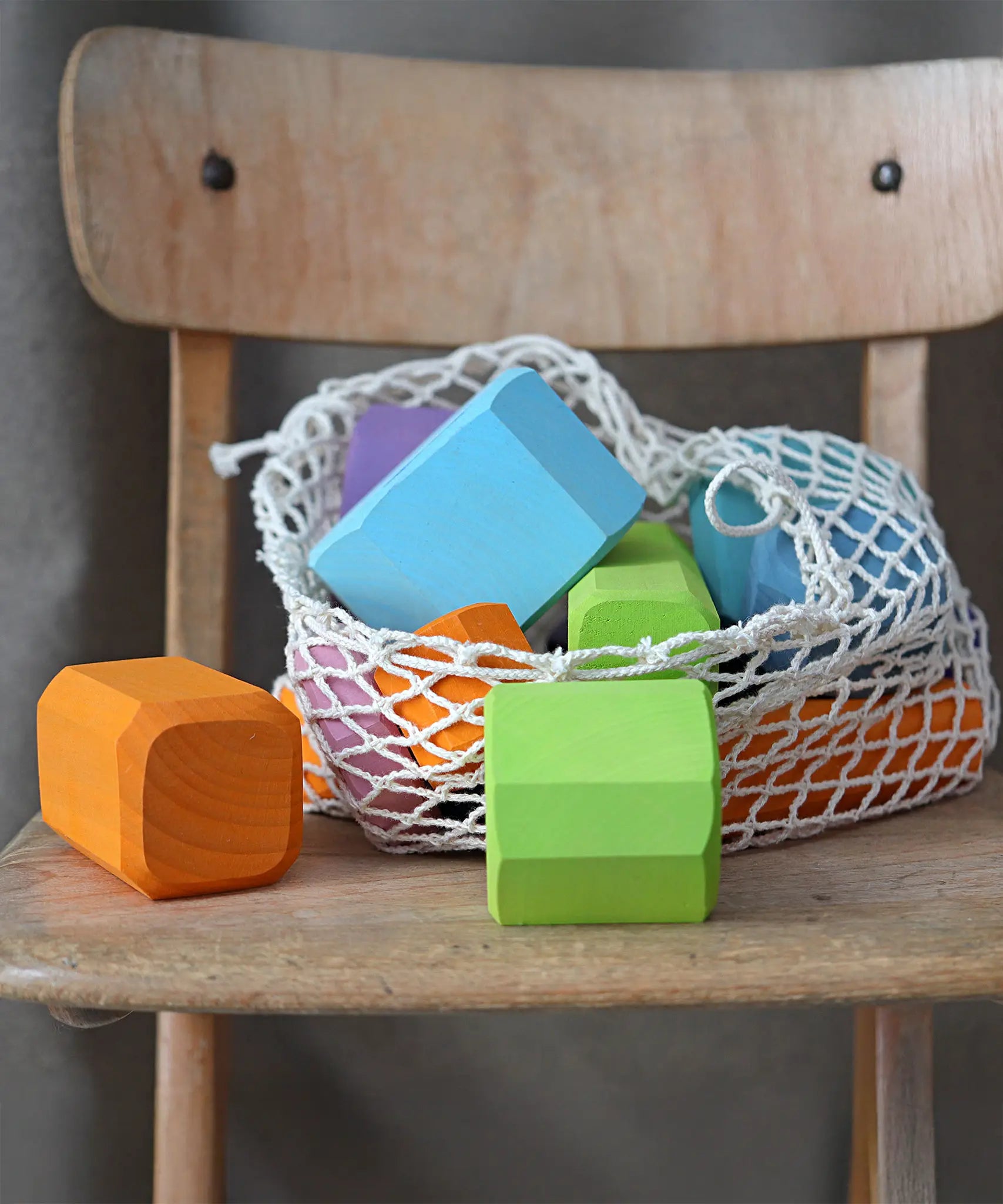 Grimm's tree slices wooden coloured building blocks shown in their netting storage bag placed on a wooden chair. 