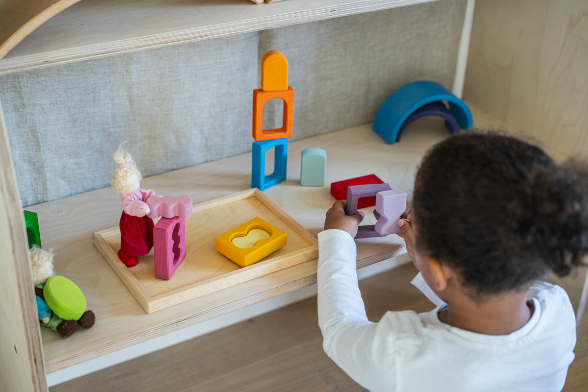 A child playing with the Grimm's Windows Building & Stacking Set. The child is holding the purple coloured window piece in their hand taking the shaped inset out of the window frame. These Grimm's toys are part of a wide range of wooden toys available here at Babipur. 