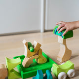 A close up of a child's hand playing with the Grimm's green squirrel Building Set. Wooden blocks in various shades of green and natural wood in a green coloured wooden storage tray. These Grimm's toys are part of a wide range of wooden toys available here at Babipur. 