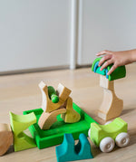 A close up of a child's hand playing with the Grimm's green squirrel Building Set. Wooden blocks in various shades of green and natural wood in a green coloured wooden storage tray. These Grimm's toys are part of a wide range of wooden toys available here at Babipur. 