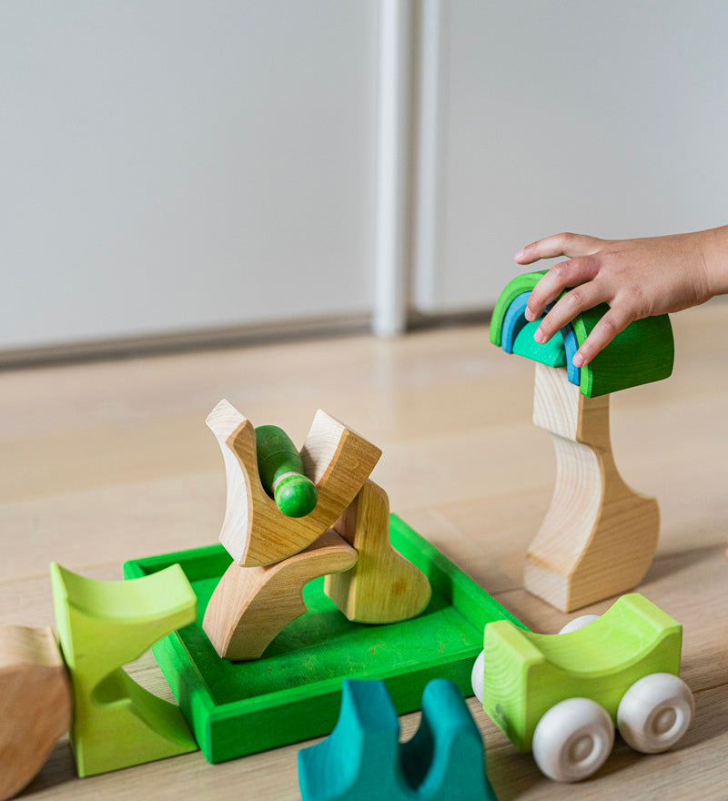A close up of a child's hand playing with the Grimm's green squirrel Building Set. Wooden blocks in various shades of green and natural wood in a green coloured wooden storage tray. These Grimm's toys are part of a wide range of wooden toys available here at Babipur. 