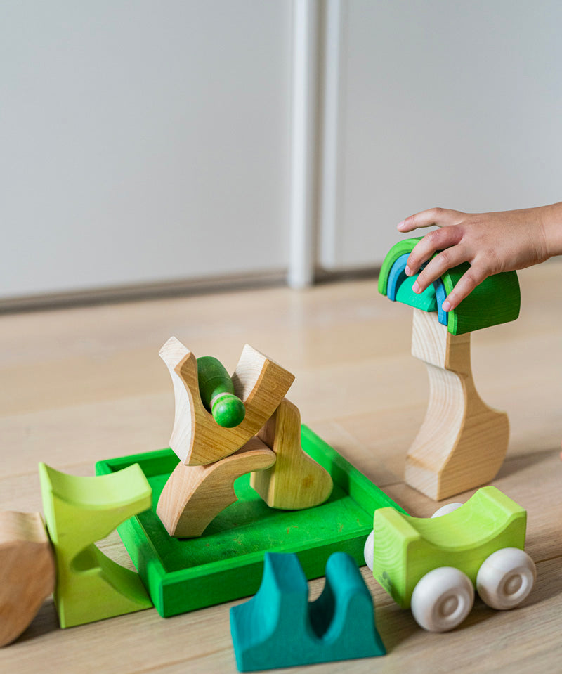 A close up of a child's hand playing with the Grimm's green squirrel Building Set. Wooden blocks in various shades of green and natural wood in a green coloured wooden storage tray. These Grimm's toys are part of a wide range of wooden toys available here at Babipur. 